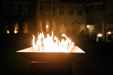 Event 4 Flamme in einer modernen Feuerschale, umgeben von historischen Gebäuden im Dunkeln.