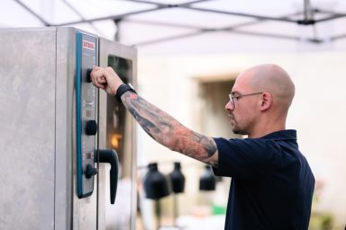Outdoorküche Ein Mann mit Tätowierungen bedient einen Backofen unter einem Zelt.
