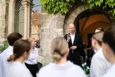 Briefing Ein Mann spricht zu einer Gruppe junger Menschen vor einem historischen Eingang.