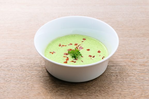 Grüne Suppe in einer weißen Schüssel, garniert mit roten Pfefferflocken und Kräutern.