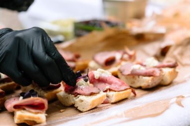 Canapes 3 Eine Hand in schwarzer Handschuh legt Belag auf kleine Brötchen mit Aufschnitt.